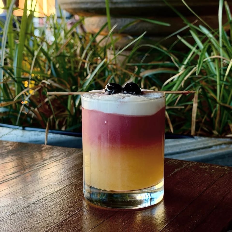 Layered pink and golden cocktail topped with white foam, black cherries, and a wooden pick on a restaurant table.