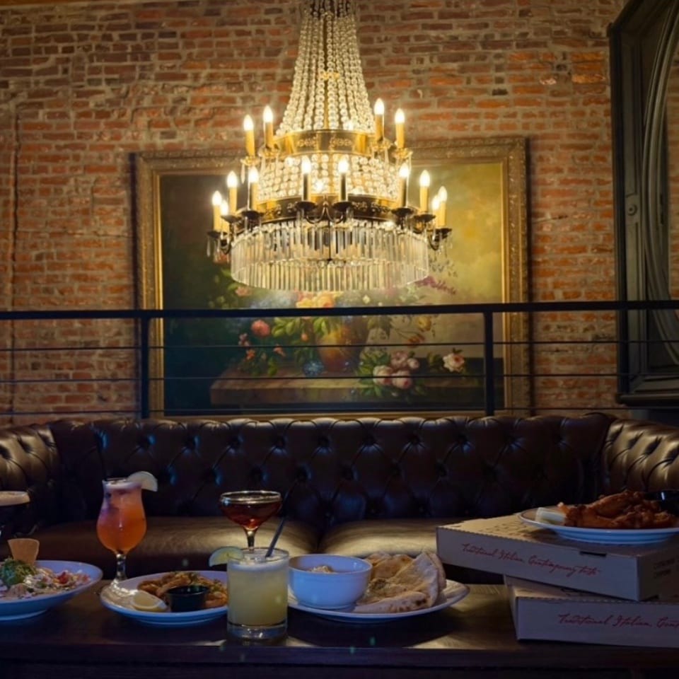 Restaurant lounge with a crystal chandelier, exposed brick walls, leather booth seating, cocktails, and plates of food on a table.