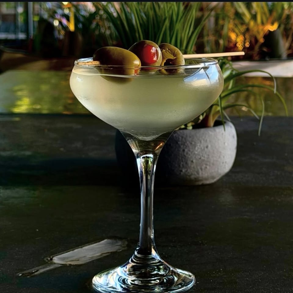 Cloudy cocktail in a coupe glass garnished with green and red olives, set on a dark table beside potted greenery.