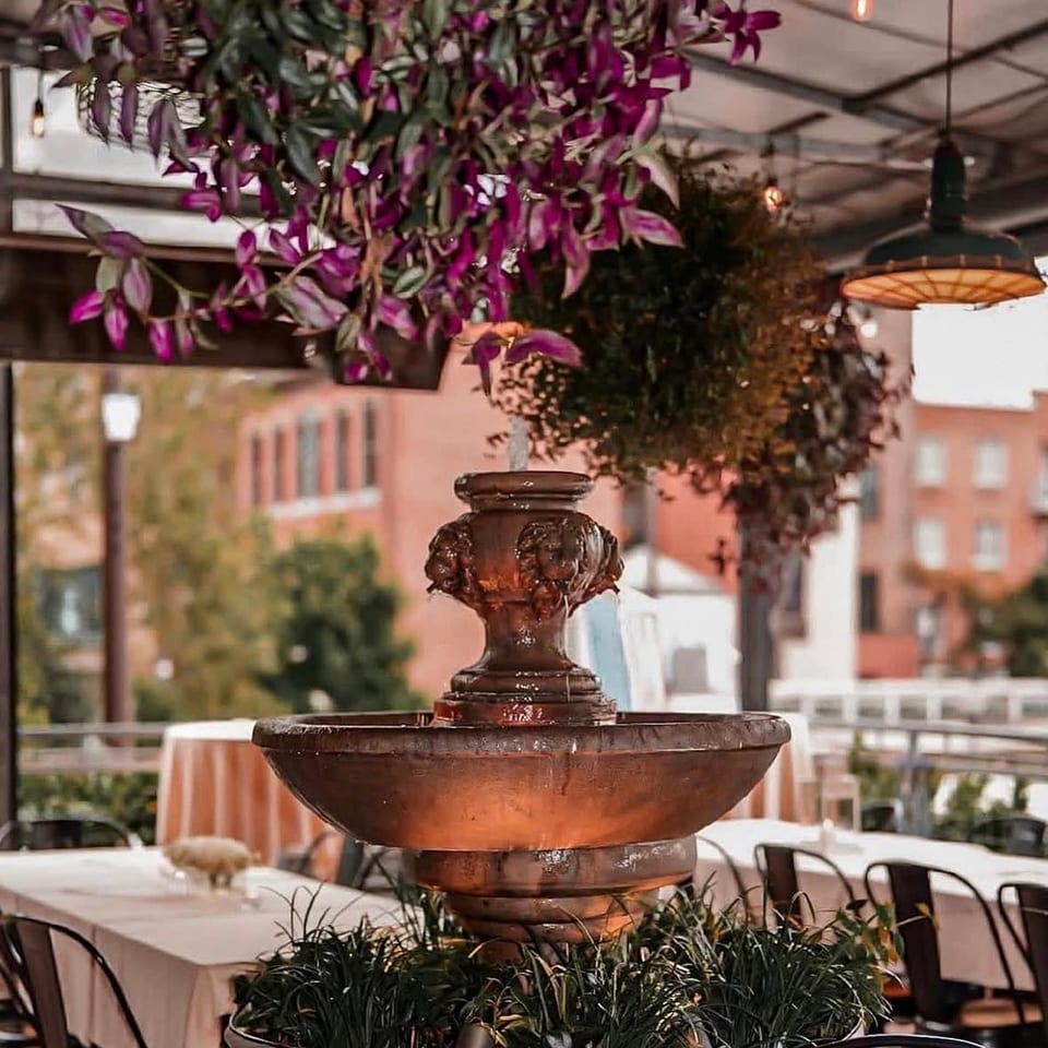 Ornate tiered fountain surrounded by greenery in a bright restaurant dining area with hanging plants and pendant lights.