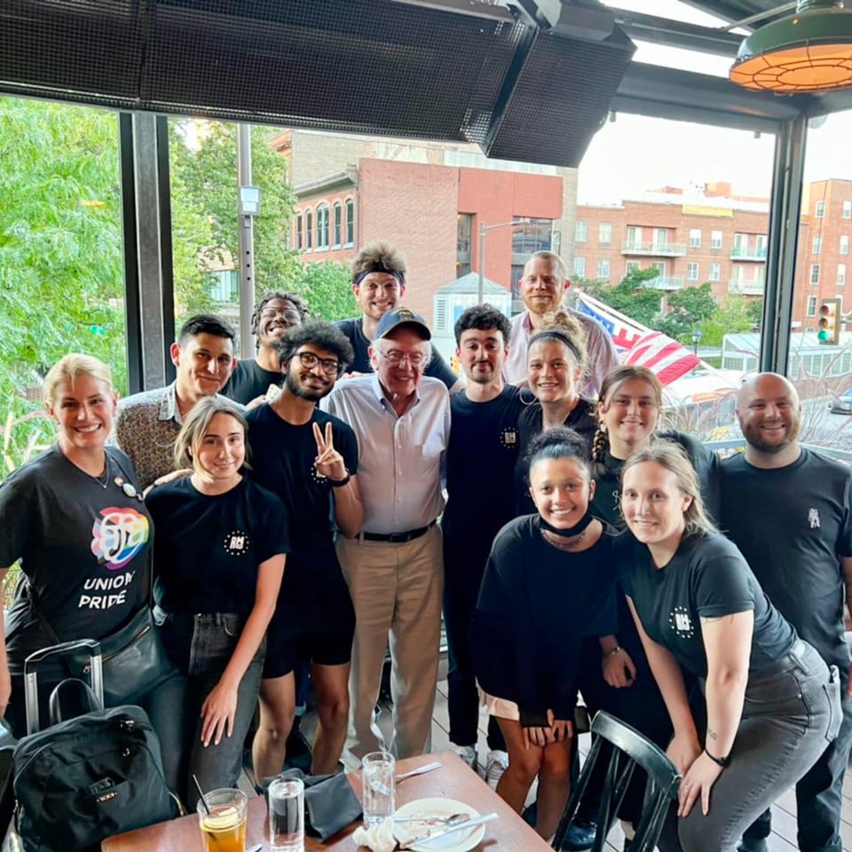Bernie Sanders poses with a group of smiling Revolution House staff on an outdoor patio.