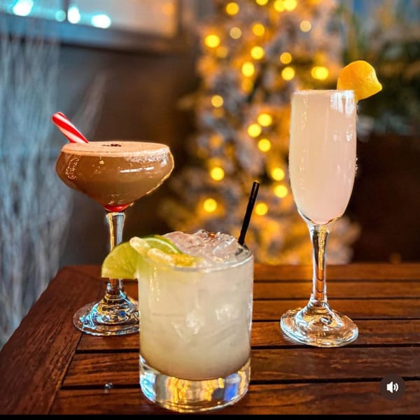 Three festive cocktails, including a peppermint martini, on a wooden table with blurred holiday lights.
