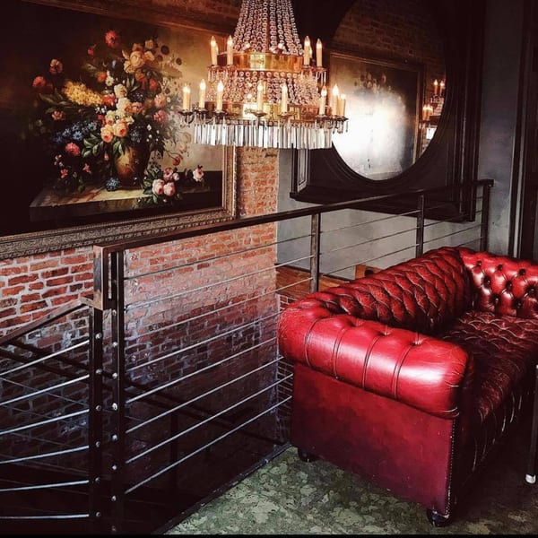 A red tufted leather sofa next to a railing, brick wall, and a large floral painting under a chandelier.