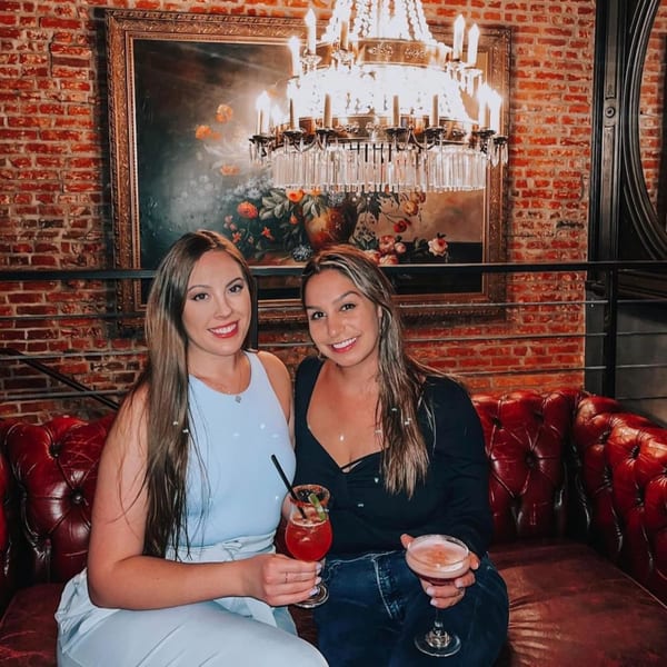 Two women holding cocktails, smiling on a red leather couch at Revolution House.