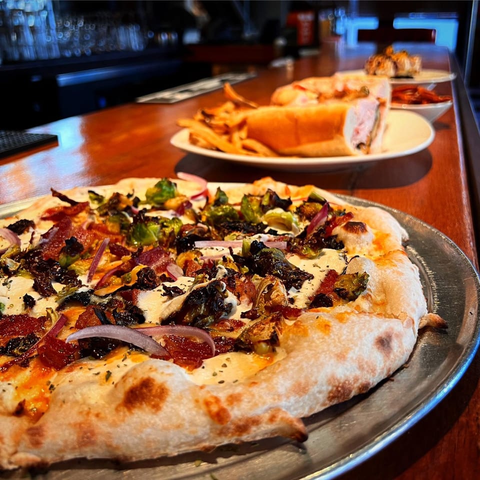 A Revolution House pizza with brussels sprouts, bacon, and red onion, with a sandwich and fries in the background.