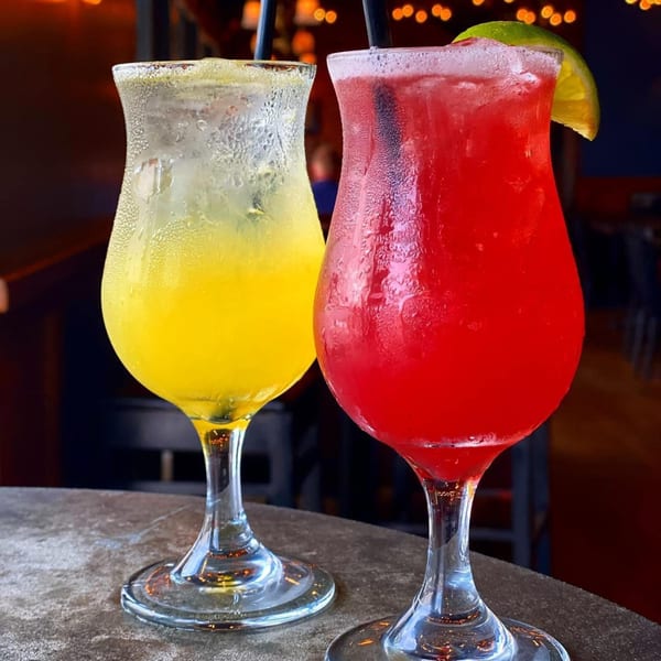 Two vibrant cocktails, one yellow and one red, in tall glasses with straws and a lime garnish.