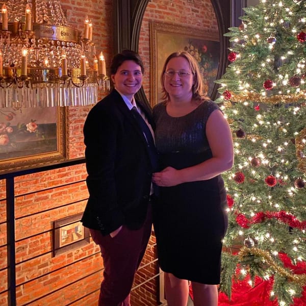 Two smiling women stand next to a decorated Christmas tree and a chandelier in Revolution House.