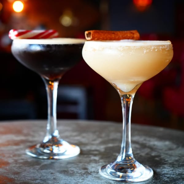 Two cocktails, one dark with a candy cane, one light with a cinnamon stick, on a table.